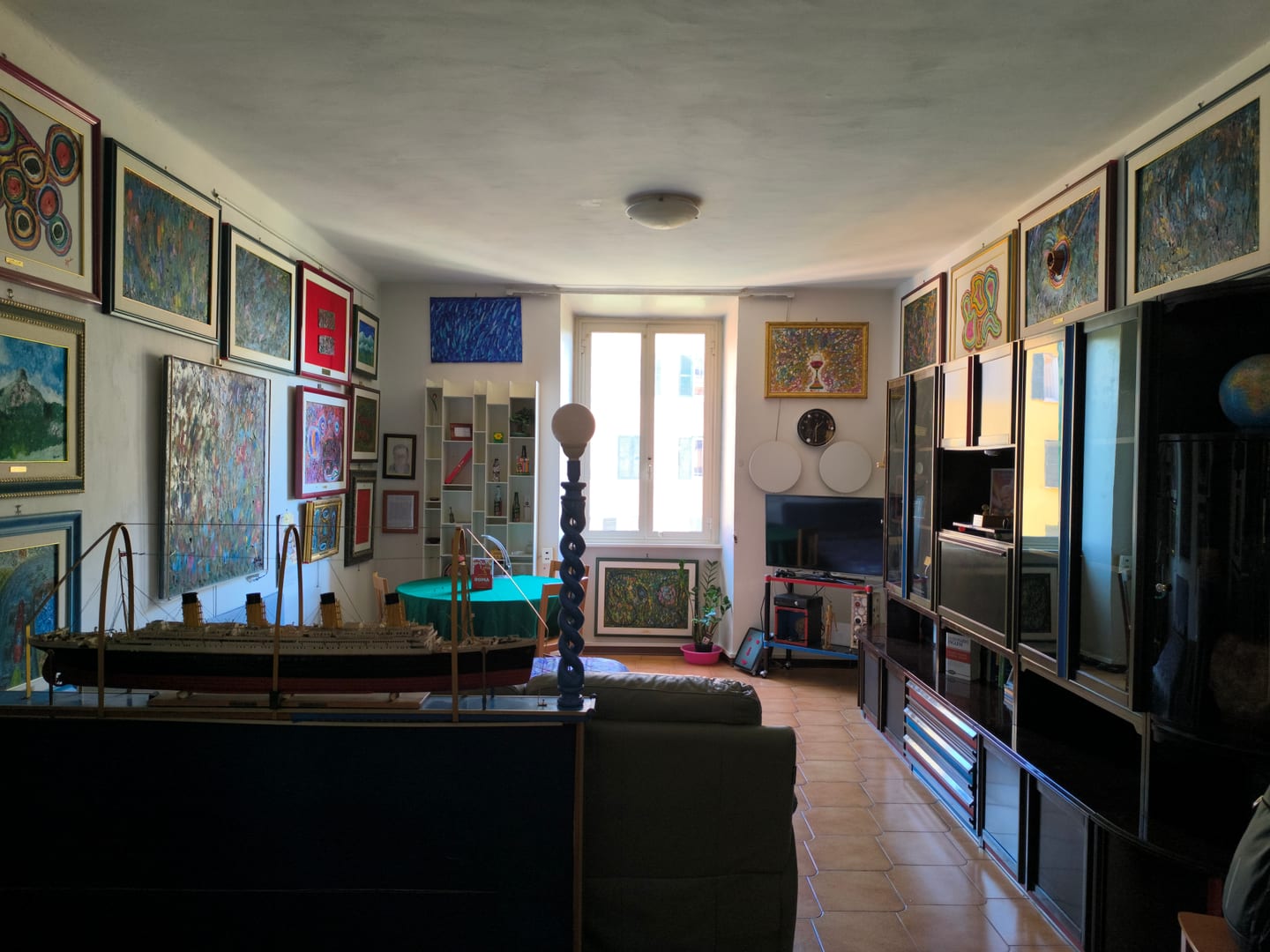 The living room seen lengthwise, with framed paintings covering both walls, a model ship on a blue plinth and a green baize table by the window